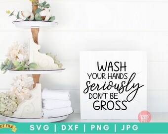 May include: White sign with black text that reads "Wash your hands seriously don't be gross". The sign is sitting on a white shelf with a tiered tray behind it. The tray is decorated with white flowers and white towels.