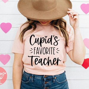May include: A pink t-shirt with the text "Cupid's Favorite Teacher" in black script font.