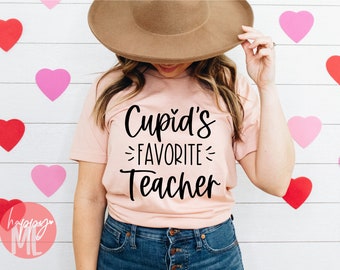 以下が含まれることがあります： 黒い筆記体で「Cupid's Favorite Teacher」と書かれたピンクのTシャツです。