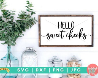 May include: A white sign with a brown wooden frame, featuring the text "HELLO sweet cheeks" in black script font. The sign is displayed on a white wall with greenery in a vase to the left.