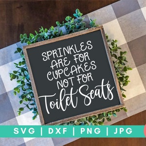 K&ouml;nnte beinhalten: Ein schwarzes Schild mit wei&szlig;em Text, auf dem steht "Sprinkles are for cupcakes not for toilet seats". Das Schild ist in Holz gerahmt und von gr&uuml;nem Laub umgeben.