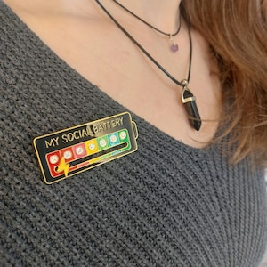 Könnte beinhalten: Eine goldene und schwarze Emaille-Anstecknadel in Form einer Batterie mit dem Text "MY SOCIAL BATTERY" und einer Reihe von Emojis, die verschiedene Glücksstufen darstellen. Die Anstecknadel wird auf einem grauen Pullover getragen.