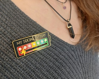 May include: A gold and black enamel pin shaped like a battery with the text "MY SOCIAL BATTERY" and a series of emojis depicting different levels of happiness. The pin is worn on a gray sweater.