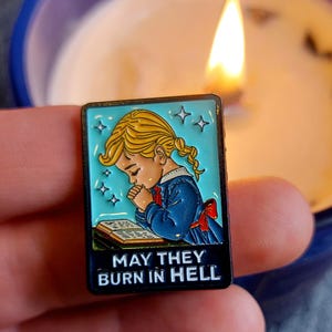 Könnte beinhalten: Rechteckiger Emaille-Pin mit einem Cartoon-Kind in einem blauen Kleid, das über einem Buch betet. Der Pin hat einen türkisfarbenen Hintergrund mit Sternakzenten und dem Text "MAY THEY BURN IN HELL" in Weiß.