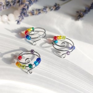 Könnte beinhalten: Drei handgefertigte Drahtringe mit bunten Perlen. Die silberfarbenen Ringe haben ein Spiraldesign mit einer Schlaufe am Ende. Die Perlen sind in einem Farbverlauf angeordnet und bilden ein auffälliges Accessoire.