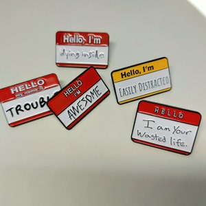Könnte beinhalten: Fünf Emaille-Pins mit rotem und gelbem Hintergrund und weißem Text. Die Pins sagen "Hello, I'm dying inside", "Hello, I'm Awesome", "Hello, I'm Easily Distracted", "Hello my name is Trouble", and "Hello I am Your Wasted Life."