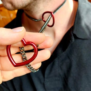Könnte beinhalten: Eine silberne Kettenkette mit einem roten herzförmigen Anhänger. Die Kette ist an einem roten herzförmigen Ring befestigt.