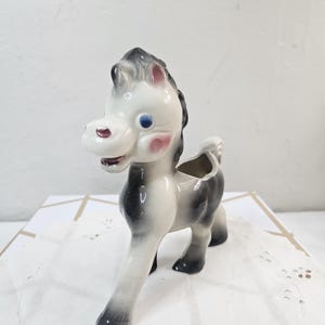 May include: A ceramic horse planter in a vintage style. The horse is primarily white with black and gray accents, including its mane and legs. It has blue eyes, pink cheeks, and a red mouth. The planter has a hollowed-out back for holding small plants or flowers.