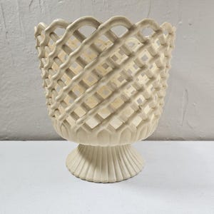 May include: Off-white ceramic basket-style vase with a lattice design. The vase has a pedestal base with vertical ridges and a flared rim. The openwork design allows light to pass through, creating a decorative effect. The vase is approximately 7 inches tall.