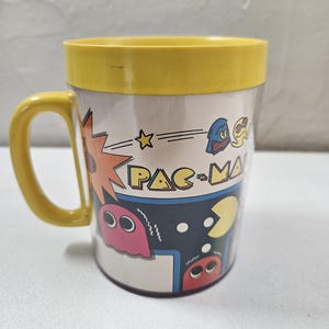May include: A yellow-handled mug featuring a Pac-Man design. The mug has a yellow rim and a wraparound graphic with Pac-Man characters and the game's logo. The design includes a pink ghost, a blue ghost, and Pac-Man himself.