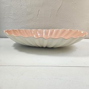 May include: A decorative, oval-shaped serving bowl with a scalloped edge. The interior is a soft pink, contrasting with the off-white exterior. The bowl is made of ceramic and is suitable for serving food.