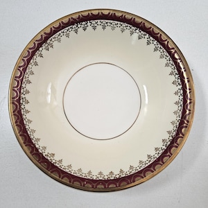 May include: A white saucer with a gold and burgundy rim. The rim has a decorative pattern of gold and black.