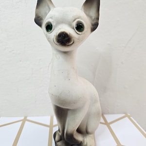 May include: A ceramic chihuahua figurine in a seated pose. The dog has a white body with dark gray ears and feet. It features green eyes and a black nose. The figurine is placed on a geometric patterned surface.