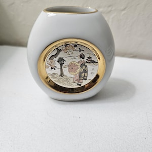 May include: White ceramic vase with a gold rim and a round medallion featuring a Japanese woman in a traditional kimono holding a fan. The medallion is surrounded by a gold border and has a floral design.