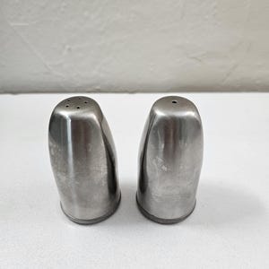 May include: Two stainless steel salt and pepper shakers. The shakers are silver, with a rounded shape and a textured base. One shaker has multiple holes on top, while the other has a single hole.