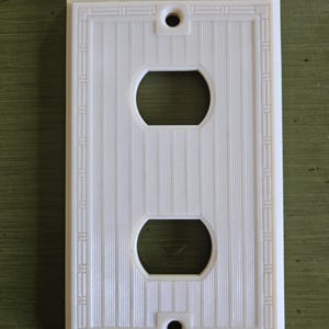 May include: A rectangular white plastic electrical outlet wall plate featuring a vertical pinstripe texture and a decorative border. The plate includes two circular openings for a duplex receptacle and two screw holes at the top and bottom.
