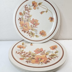 May include: A stack of beige ceramic plates with a brown rim and floral design. The plates feature orange and yellow flowers with green leaves. The plates are stacked on a white surface.