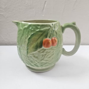 May include: A green ceramic pitcher with a leaf and cherry design. The pitcher has a handle and a spout for pouring. The cherries are orange and the leaf is green. The pitcher is likely for serving beverages.