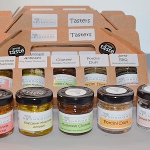 May include: A selection of mushroom-based products in small glass jars with black lids, displayed in a cardboard carrier. The jars contain various mushroom preparations, including pickles, chutney, and dust. Labels identify each flavour.