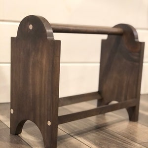 May include: A dark brown wooden magazine rack with a horizontal bar at the top. The rack has a rectangular base and arched side supports. Small silver-coloured rivets are visible on the sides.
