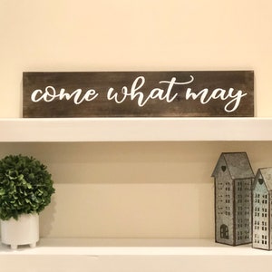 May include: A wooden sign with the text "come what may" in white paint. The sign is on a white shelf with a small green bush in a white pot and two metal houses on the right.