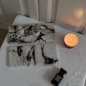May include: Two rectangular marble trays with black veining, holding perfume bottles. A lit candle in a red container and a black perfume bottle are also on a white surface. The Tom Ford Oud Wood perfume bottle is visible.