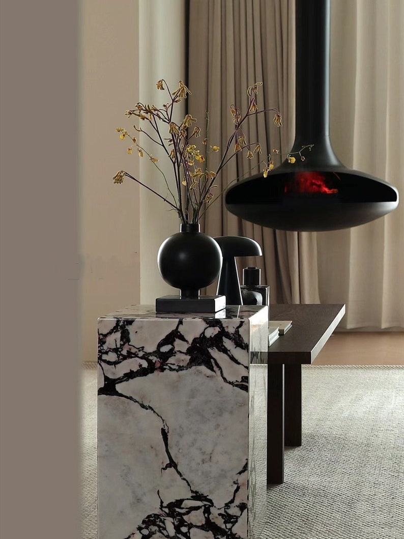 May include: A black marble pedestal with a black vase holding dried yellow flowers. A black hanging fireplace with a glowing fire is in the background.