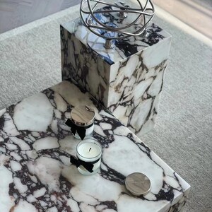 May include: Two white marble square tables with black veining. The top table has two candles with black ribbon and a silver lid. The bottom table has a silver metal orb sculpture on top.