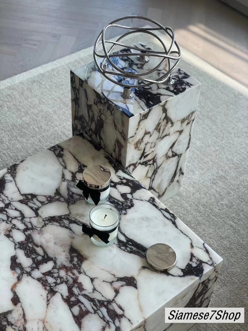 May include: Two white marble square tables with black veining. The top table has two candles with black ribbon and a silver lid. The bottom table has a silver metal orb sculpture on top.