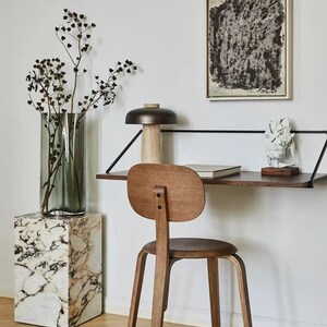 May include: A wooden chair with a rounded back and seat sits in front of a floating desk with a black metal frame. A lamp with a brown shade and a wooden base sits on the desk. A vase with dried branches sits on a white marble pedestal.