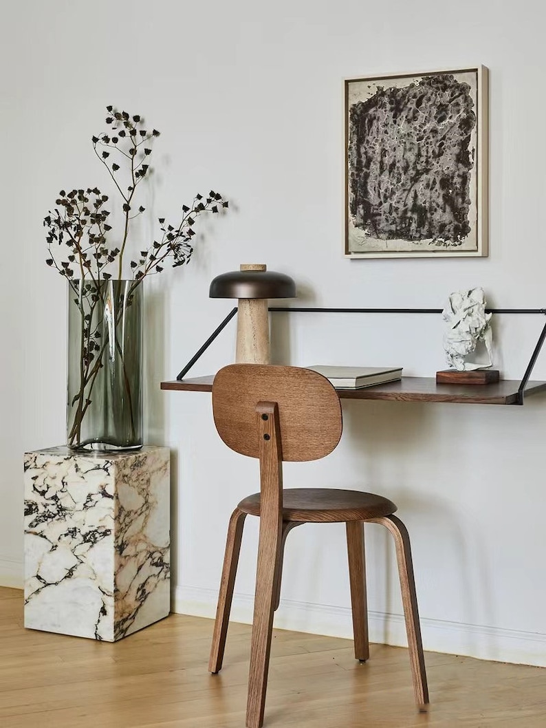 May include: A wooden chair with a rounded back and seat sits in front of a floating desk with a black metal frame. A lamp with a brown shade and a wooden base sits on the desk. A vase with dried branches sits on a white marble pedestal.