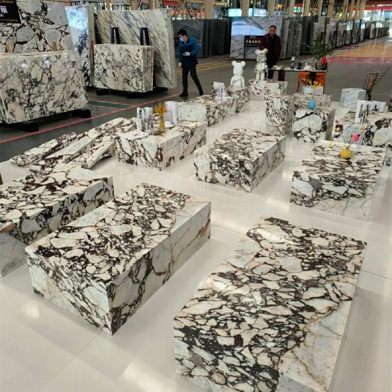 May include: A showroom display of white marble slabs with black and brown veining. The slabs are arranged in a variety of shapes and sizes, creating a unique and modern design. The slabs are used to create a series of tables and benches, perfect for showcasing the beauty of the marble.