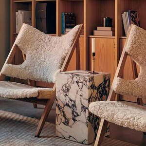 May include: Two white sheepskin chairs with wooden frames and a white marble side table in a room with a wooden bookshelf filled with books.