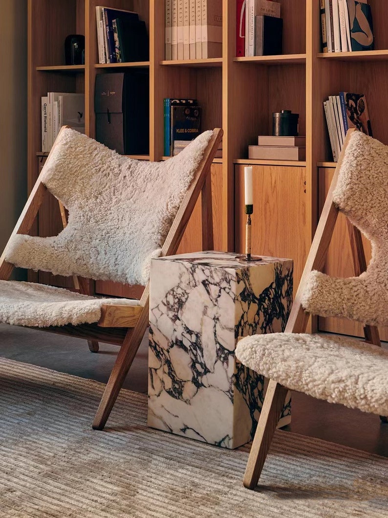 May include: Two white sheepskin chairs with wooden frames and a white marble side table in a room with a wooden bookshelf filled with books.