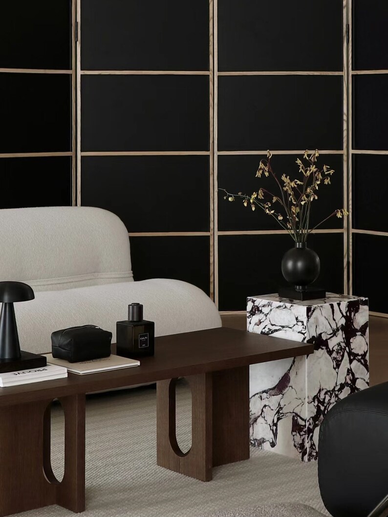 May include: A dark wood coffee table with a white couch and a black screen in the background. The table has a unique design with oval cutouts in the legs. A black vase with dried flowers sits on a white marble pedestal.