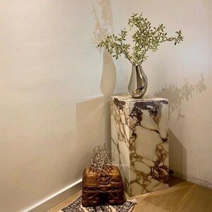 May include: A white marble pedestal with a silver vase filled with green leafy branches. A wooden box with intricate carvings sits on the floor in front of the pedestal.