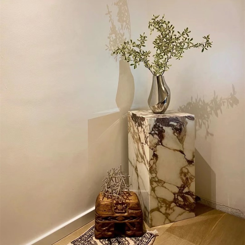 May include: A white marble pedestal with a silver vase filled with green leafy branches. A wooden box with intricate carvings sits on the floor in front of the pedestal.