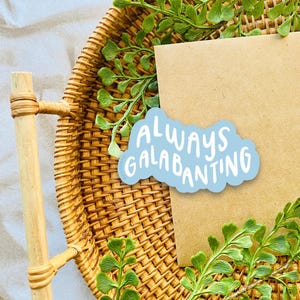May include: A woven wicker tray with a wooden handle holds a brown card and a blue sticker with the words "ALWAYS GALABANTING" in white. Green leafy sprigs are arranged around the card and tray.