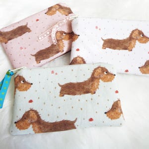 Könnte beinhalten: Drei Reißverschlusstaschen mit einem sich wiederholenden Dackel-Muster. Die Taschen sind in Rosa, Weiß und Hellblau gehalten, jede mit einem anderen Hintergrundmuster aus kleinen Punkten. Die Taschen haben Reißverschlussanhänger aus Band oder Stoff.