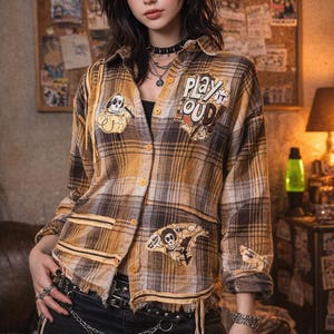 May include: A plaid button-up shirt in shades of brown and yellow, featuring patches with cartoon skull designs and the text "PLAY LOUD". The shirt has a frayed hem and is worn with a black top and denim skirt.