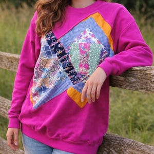 May include: A magenta sweatshirt with a patchwork design on the front. The patchwork includes various patterned fabrics in blue, floral, and geometric designs, outlined with orange and blue accents. The sweatshirt has a crew neck and long sleeves.