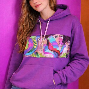 May include: A purple hoodie with a colorful graphic design featuring faces and birds. The hoodie has a kangaroo pocket and a drawstring hood. The model is wearing blue jeans. The background is a solid purple and orange.