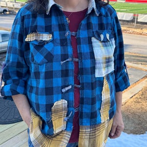 May include: A blue and black plaid flannel shirt with patches of yellow and white plaid fabric. The shirt has a button-up front and is worn over a burgundy top and blue jeans. The sleeves are rolled up, and the shirt appears to be handmade.