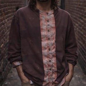 May include: A long-sleeved brown shirt with a vertical plaid panel down the center, featuring button closures. The shirt has a distressed appearance, with visible wear and tear. The sleeves are cuffed, and the shirt is worn with tan pants.