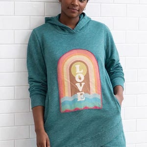 Puede incluir: Sudadera con capucha de color turquesa con un gráfico de arcoíris y la palabra "LOVE". El diseño del arcoíris presenta un sol, olas y un arco colorido. La sudadera tiene un bolsillo de canguro y un dobladillo envolvente.