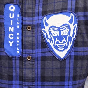 May include: A blue and black checked shirt with patches. One patch reads "Quincy Blue Devils" in white lettering on a blue background. The other patch features a white devil face on a blue background.