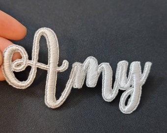 May include: A silver-colored embroidered patch featuring the name Amy in a cursive script font. The lettering has a raised, textured appearance with a satin stitch finish, creating a distinct outline for each letter in the name.