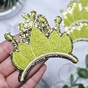 Może przedstawiać: Zielona i złota korona z brokatem o aksamitnej fakturze. Korona ma kształt półkola i spiczasty wierzchołek.