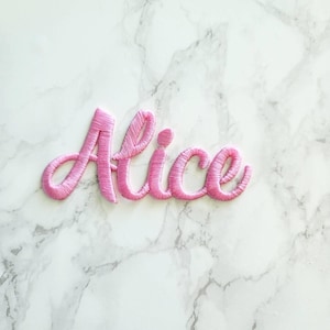 May include: A pink embroidered patch featuring the name Alice in a cursive script font. The lettering is stitched with a dense, raised thread texture, creating a distinct outline and filled appearance for each letter in the name.