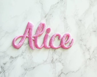 May include: A pink embroidered patch featuring the name Alice in a cursive script font. The lettering is stitched with a dense, raised thread texture, creating a distinct outline and filled appearance for each letter in the name.
